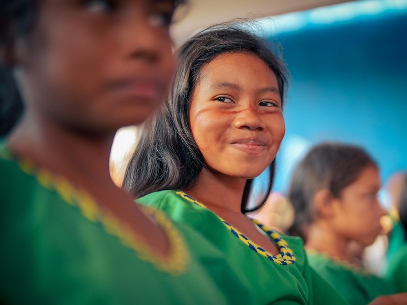 Niña indígena sonriente, mirando a la cámara en un ambiente escolar o comunitario. Al fondo se ven otras niñas con atuendos similares, ligeramente desenfocadas.